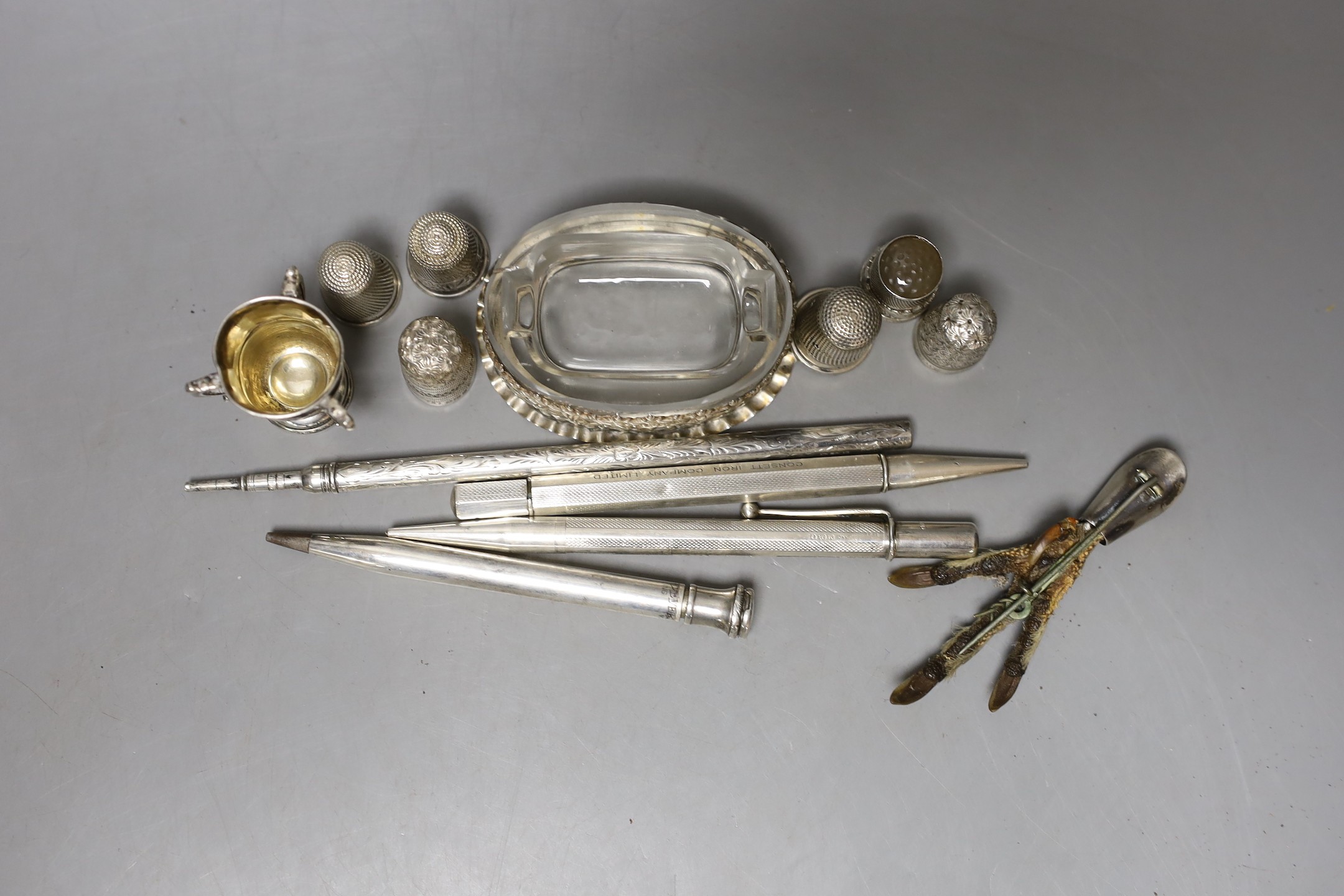 An Edwardian repousse silver mounted glass salt,69mm, a small silver tri-handled trophy cup, five silver thimbles, a plated thimble, three silver cased pens, a sterling pen, one other white metal pen and a brooch.